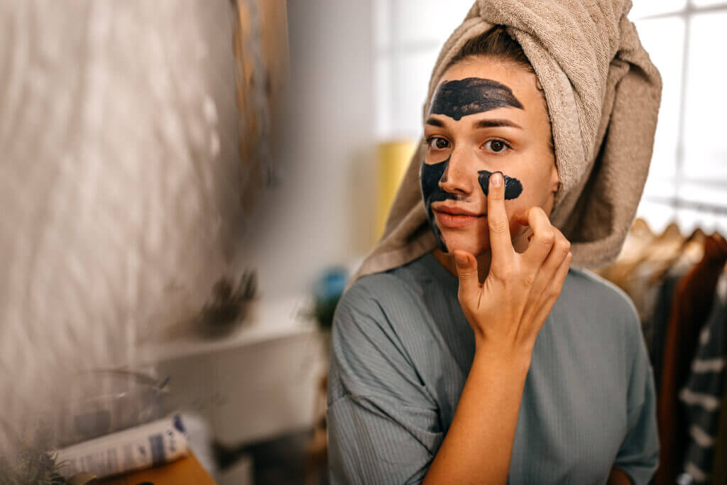 Young women with towel on head applying mineral black charcoal mask while looking at mirror