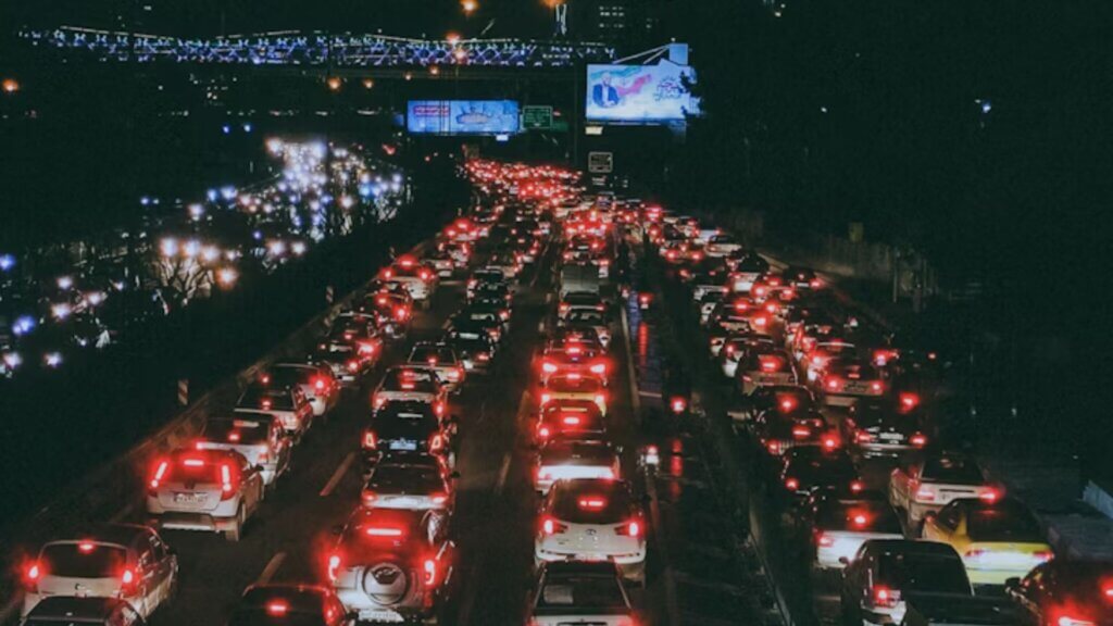 Nighttime traffic jam with rows of cars and glowing brake lights on a busy highway. Elevated bridge with lights and billboards in the background.