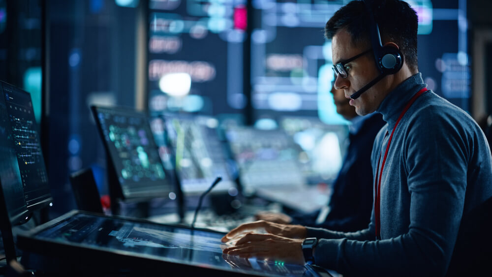 Portrait of Professional IT Technical Support Specialist Working on Computer in Monitoring Control Room with Digital Screens. Employee Wears Headphones with Mic and Talking on a Call.