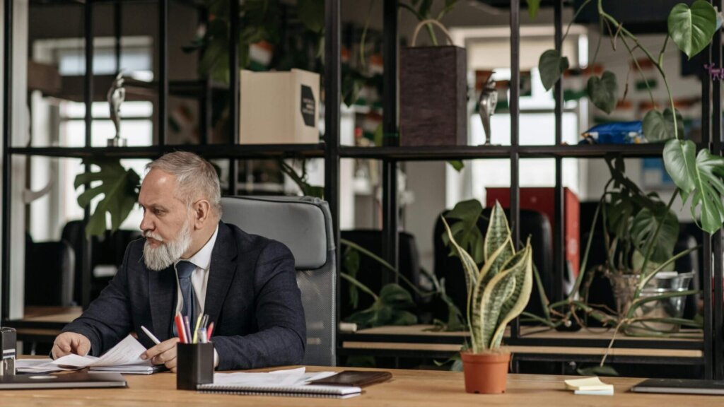 Elderly man sitting on an office chair with legs crossed.