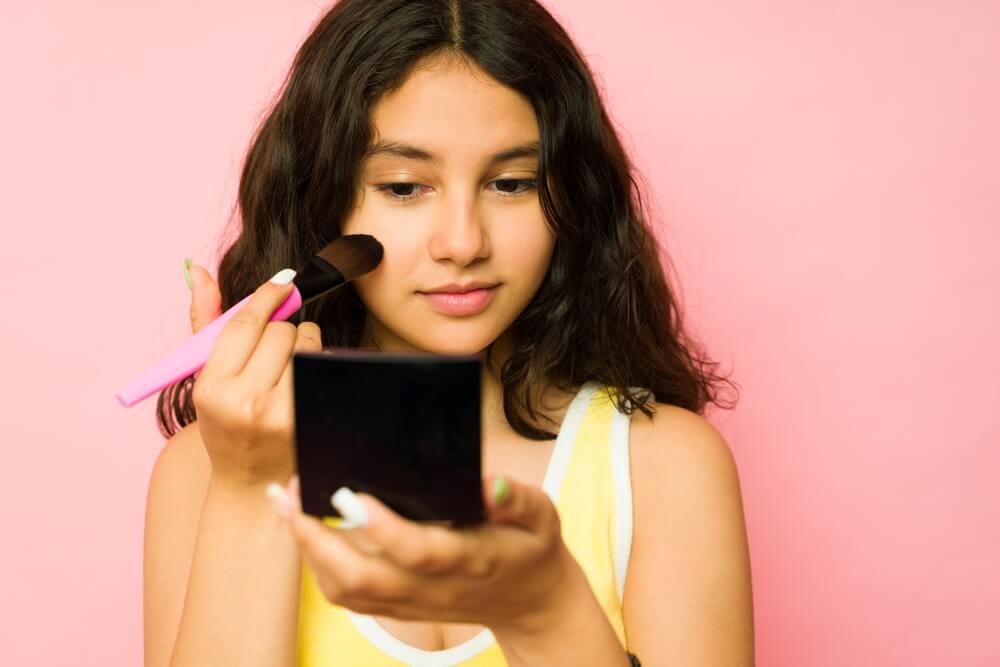 Teen girl putting on makeup blush while looking in the mirror