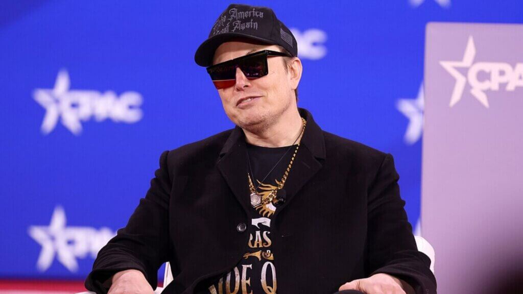 Elon Musk, wearing a black "Make America Great Again" cap, oversized sunglasses, and layered gold necklaces, sits against a CPAC backdrop while smirking.