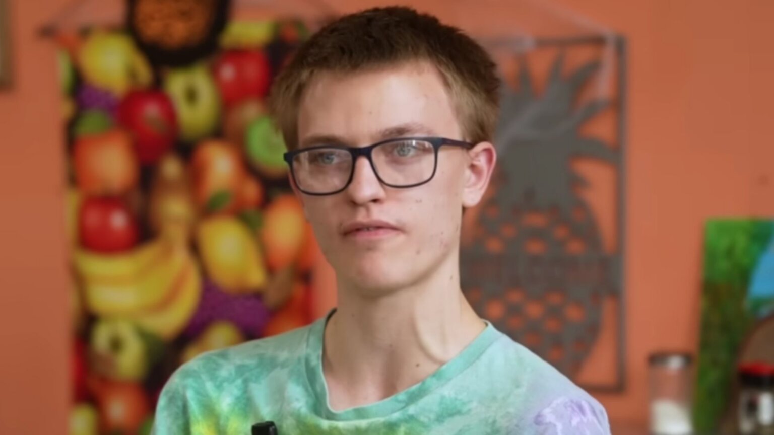 A close-up portrait of a young person wearing glasses and a tie-dye shirt, looking thoughtful while standing in a brightly decorated café interior.
