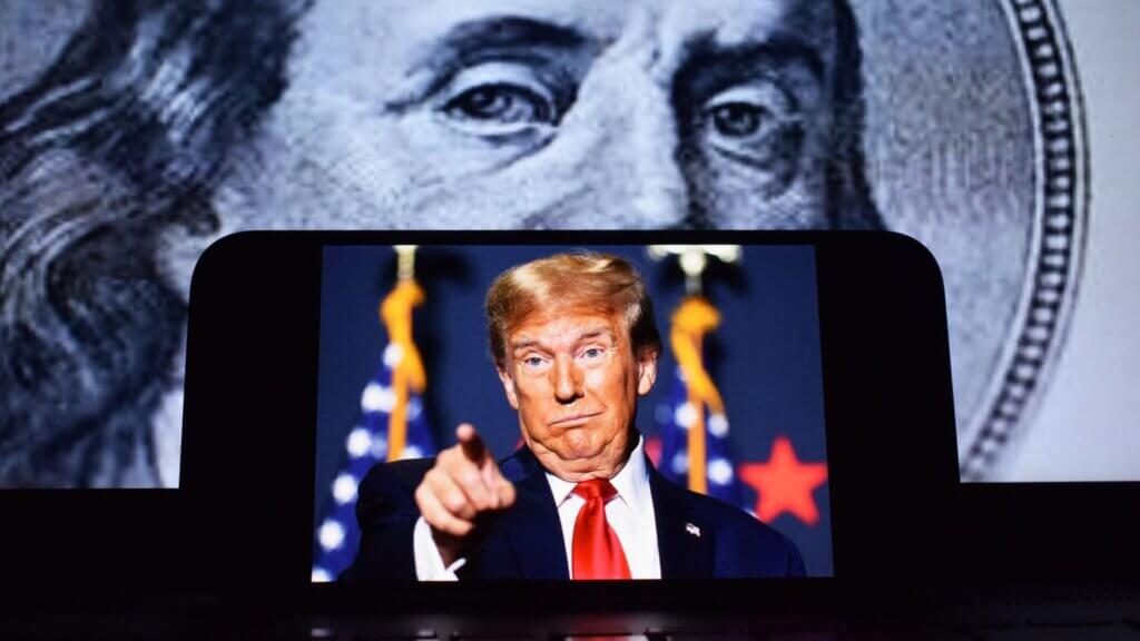 A large screen shows Pres. Donald Trump, pointing toward the camera, standing before U.S. flags, with a close-up of a U.S. hundred-dollar bill featuring a historical portrait visible in the background.