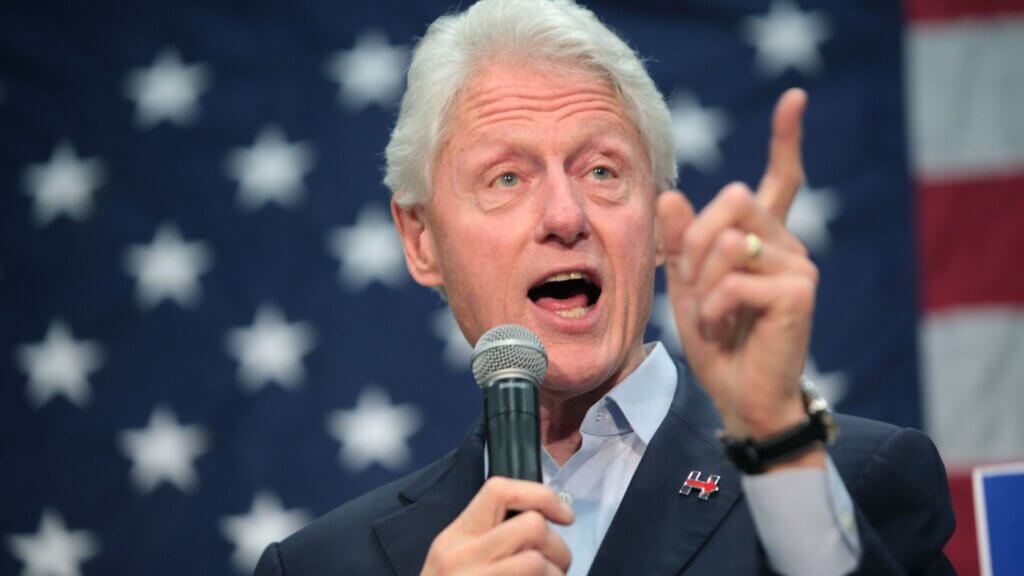 Bill Clinton speaks passionately into a microphone, pointing upward during a campaign rally with a U.S. flag backdrop, symbolizing American politics and Democratic Party engagement.