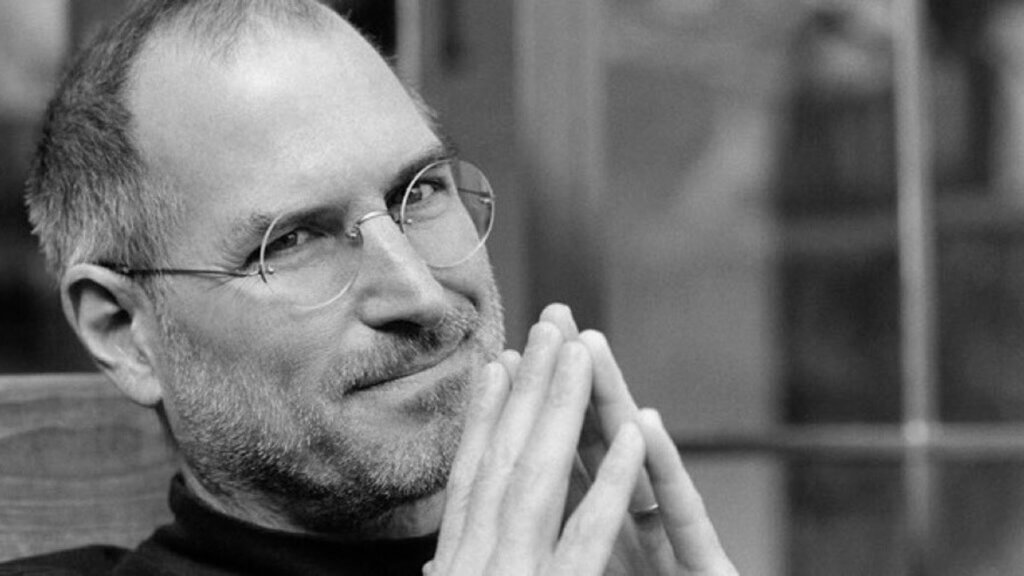 Black and white portrait of Steve Jobs smiling subtly with his hands pressed together near his face, wearing rimless glasses and a black turtleneck. A thoughtful expression hints at his visionary approach to technology and business.