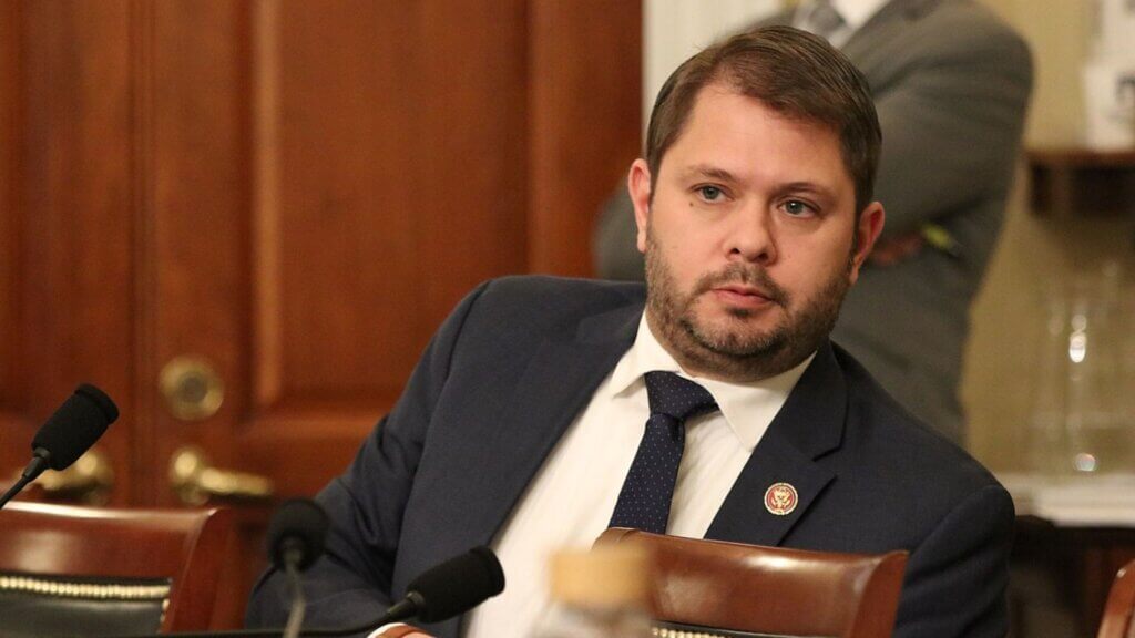 U.S. Senator Ruben Gallego sits in a congressional hearing room, wearing a dark suit and patterned tie, with a serious expression.