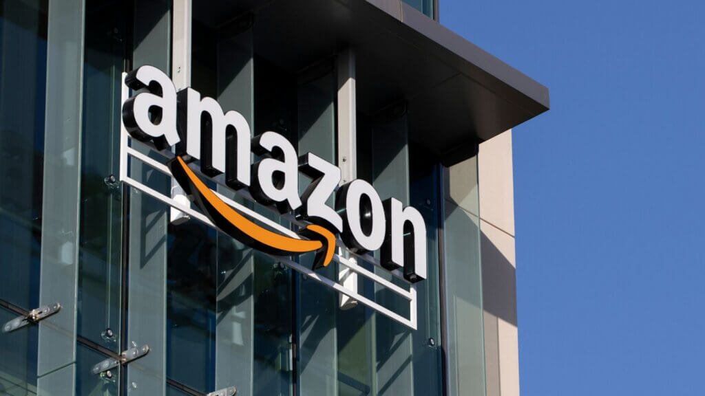 Amazon logo mounted on a glass office building with a blue sky in the background.
