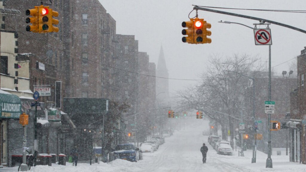 Snow-covered city street with parked cars and buildings during a winter day.