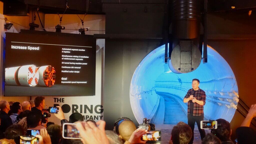 Elon Musk speaks to an audience while standing inside a large tunnel structure.
