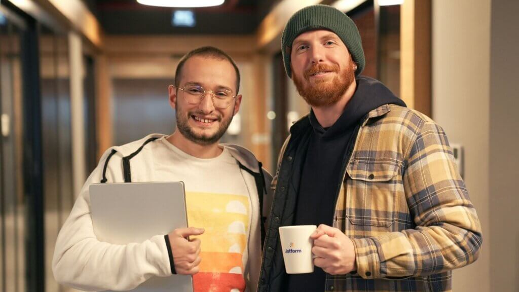Two smiling men standing in a hallway, one holding a laptop and the other a branded coffee mug.
