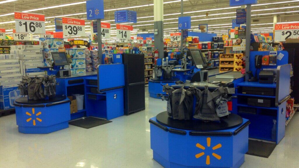 An empty Walmart checkout area with multiple self-checkout kiosks, blue counters, bagging stations, and overhead signs displaying everyday low prices.