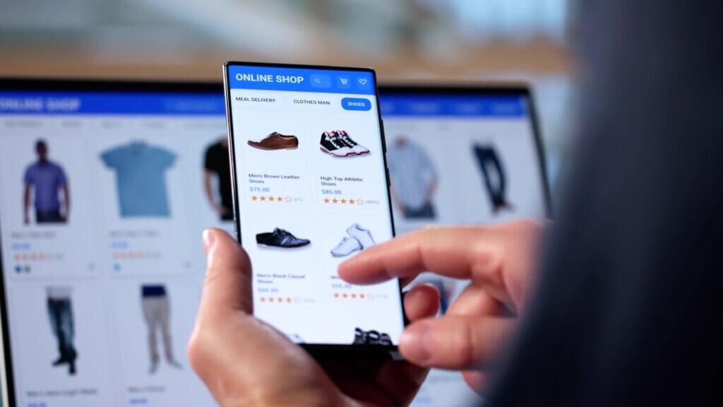 A person scrolls through an online shopping app on a smartphone, viewing men's shoes with listed prices and star ratings, with a blurred laptop screen in the background showing similar items. This image reflects consumer pricing in a digital marketplace.