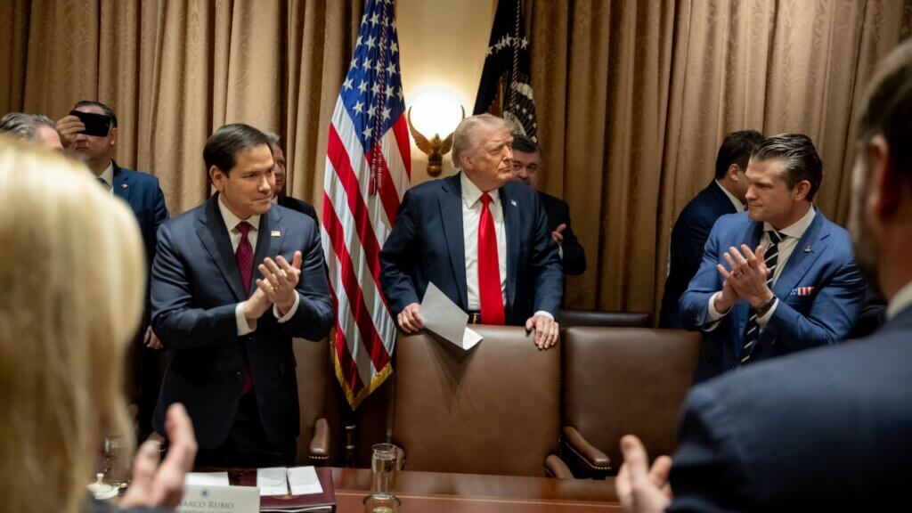 Donald Trump stands at the head of a conference table holding papers, flanked by applauding officials including Marco Rubio, with American flags and presidential emblems in the background.