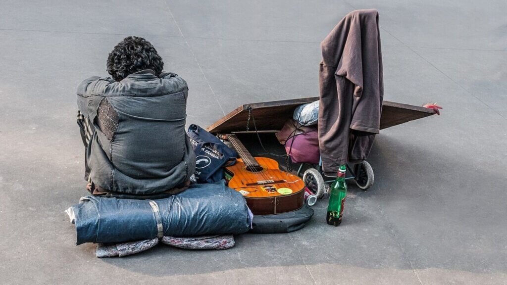 A person sits on the ground next to personal belongings and a guitar.