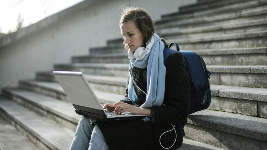 Young woman sitting on outdoor steps using a laptop, with headphones around her neck and a backpack.