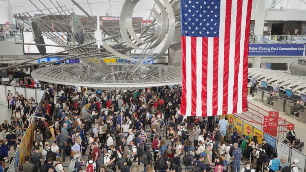 Large crowd of travelers waiting in a U.S. airport security line beneath an American flag.