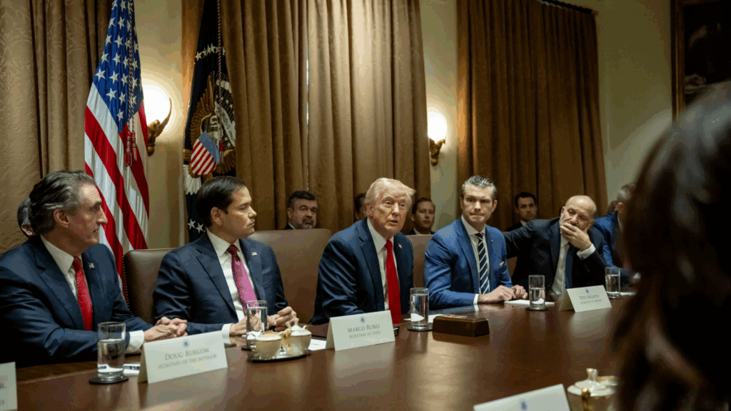 Group of government officials seated around a conference table with U.S. flags in the background.