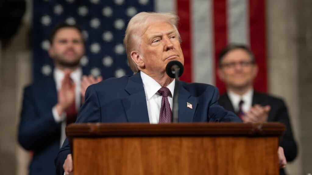 Donald Trump stands at a podium with a stern expression during a formal address, as people applaud behind him and an American flag hangs in the background, representing American politics.