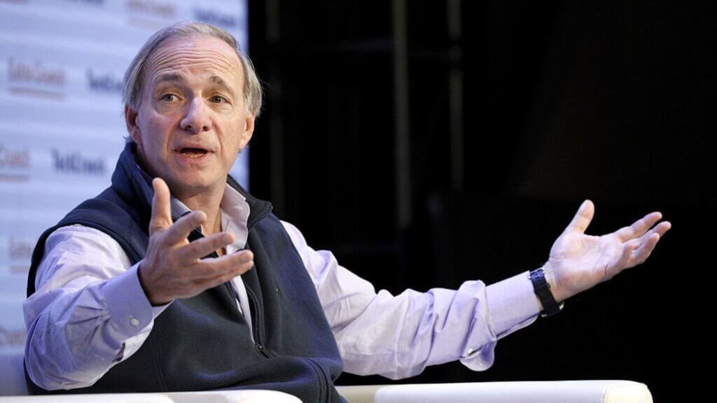 Ray Dalio speaking at an event, gesturing with open hands while seated on a white chair. The hedge fund manager is known for commenting on economic trends, including concerns when US debt hits $38 trillion.