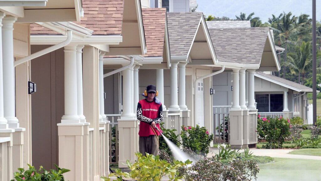 A maintenance worker in protective gear sprays landscaping in front of uniform suburban homes lined with white columns and manicured shrubs.