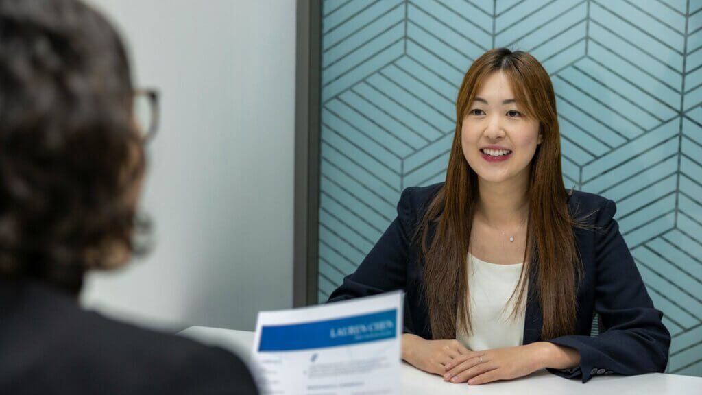 Woman in a business suit smiling during a job interview across the table from an interviewer holding a résumé.