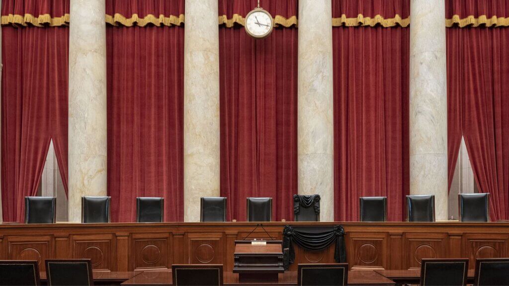 The interior of the U.S. Supreme Court chamber, featuring red curtains, marble columns, a central clock, the judge’s bench, and rows of empty seats.
