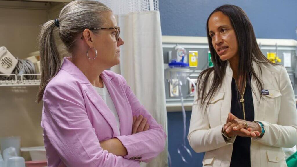 Gov. Hobbs and a staff member in conversation inside a medical facility, one wearing a pink blazer and the other a white jacket.