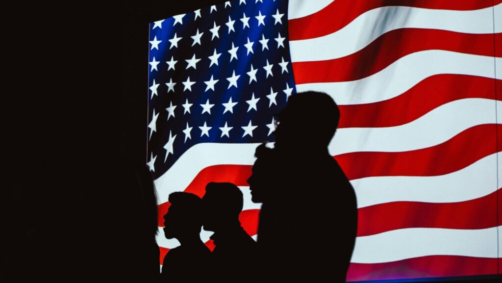 Silhouettes of four people standing in front of a large United States flag backdrop.