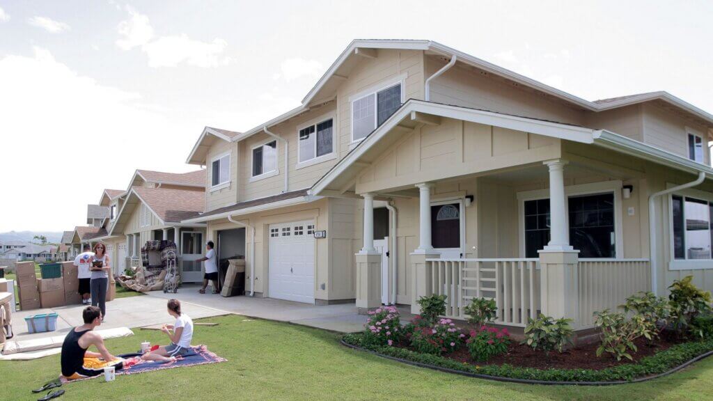 People move into beige two-story suburban homes with porches, surrounded by boxes and unpacked furniture, while two individuals have a picnic on the grass nearby.