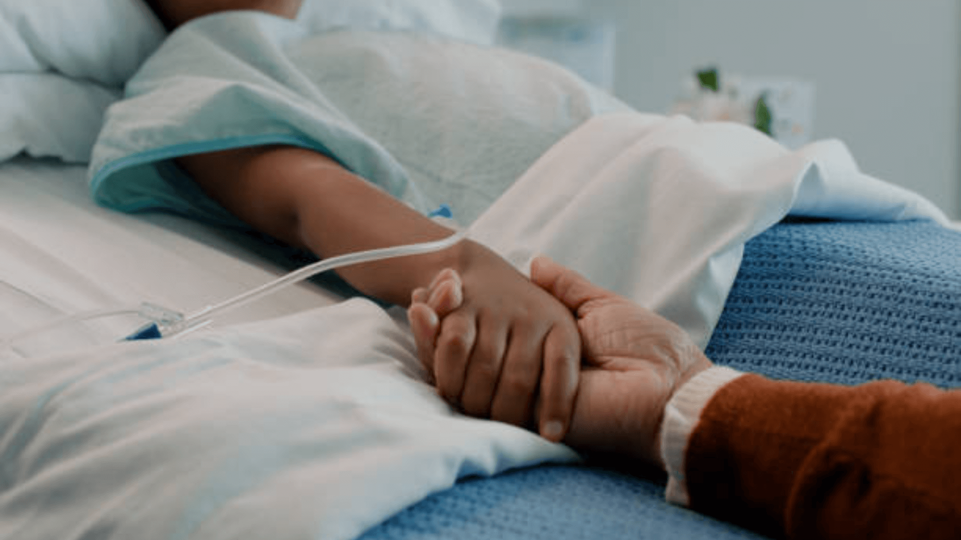 Close-up of a child’s hand in a hospital bed being held by an adult.
