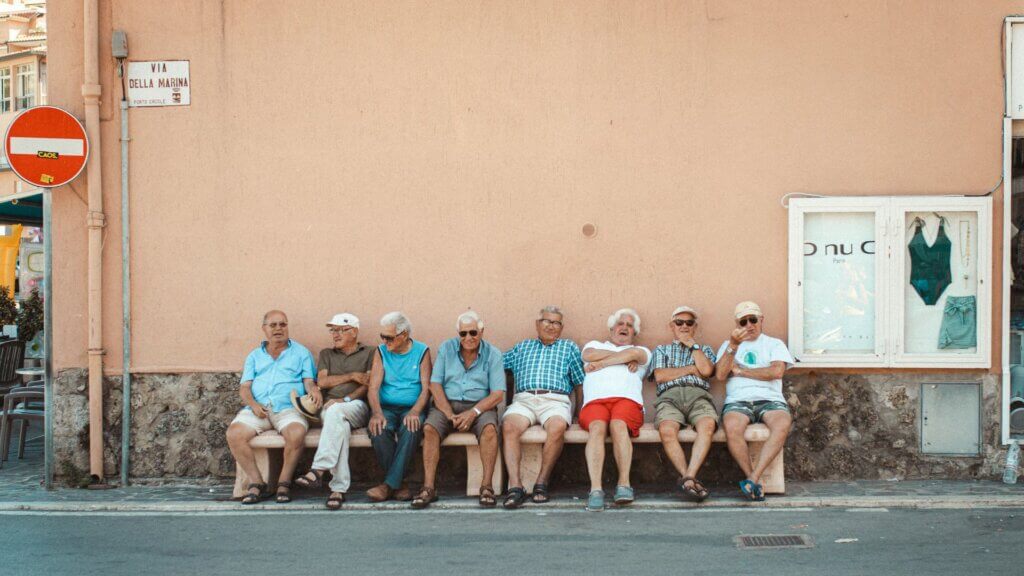 Eight men sitting on a bench against an orange wall during the day