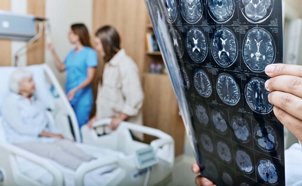 Doctor holding a patient's MRI result with the senior patient sitting upright on the hospital bed in the background, next to a relative and the nurse