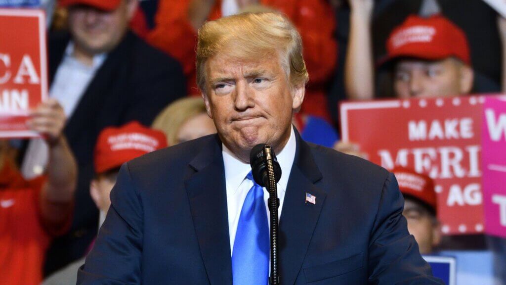 President Trump, in a dark suit and blue tie, stands at a podium during a crowded political rally, with supporters in red hats holding signs behind him.