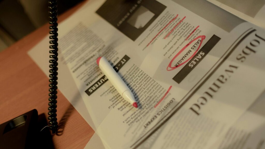 A person holding a phone while sitting at a desk with a laptop and papers, appearing to make a call during a job search.