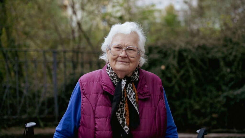 Portrait of an elderly woman with short gray hair and round glasses looking directly at the camera with a soft, thoughtful expression.