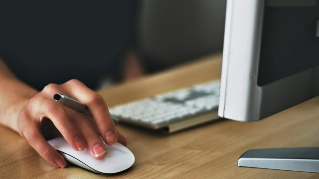 Person using a white Apple Magic Mouse on a desk while working on a computer.