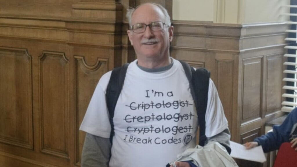 Smiling Bill Burr, wearing glasses and a backpack, stands indoors in front of wood-paneled walls. His T-shirt reads "I am a cryptologist," with several misspelled versions crossed out, followed by "I break codes."