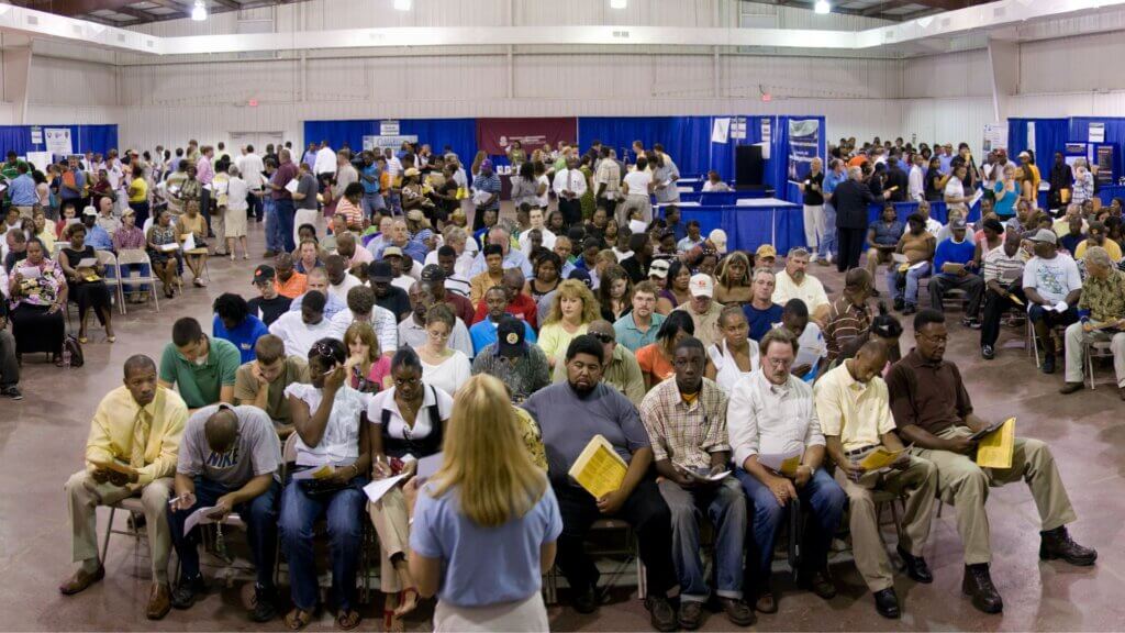 A large group of people sit closely together in a convention hall, many holding papers while listening to someone speaking at the front.