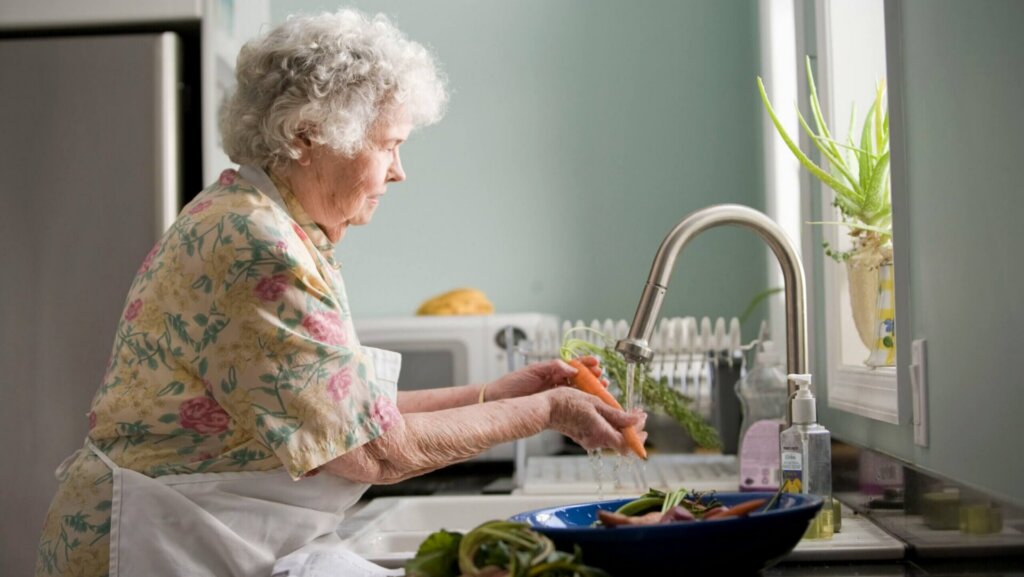 Woman wearing a yellow and pink floral dress standing outdoors, rinsing fresh carrots under running water.