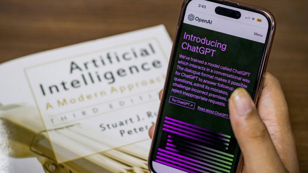 Close-up of a hand holding a smartphone showing the ChatGPT interface with a book in the background, representing artificial intelligence technology.