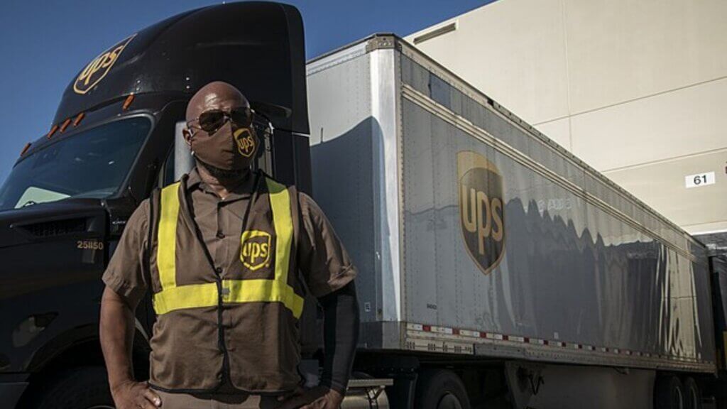 A delivery driver wearing a safety vest and face covering stands in front of a UPS delivery truck outside a warehouse.