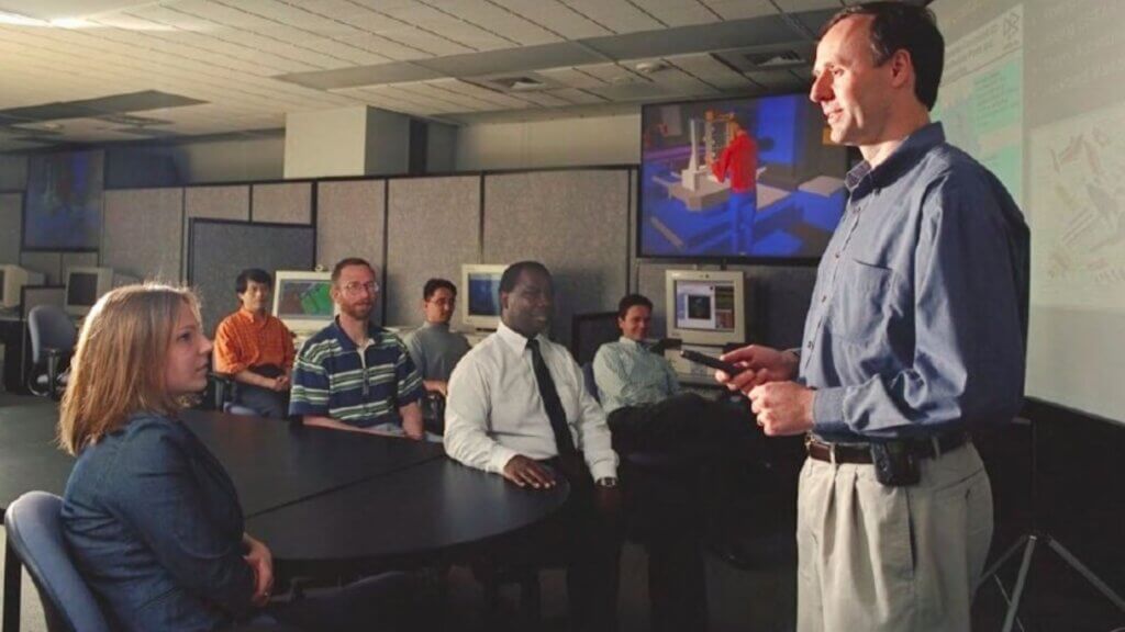 Man stands at the front of NIST conference room giving a presentation to a small group seated at a table with desktop computers behind them.