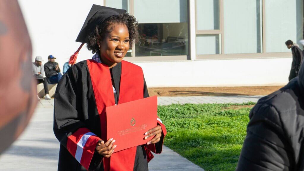Woman in graduation gown holding a red diploma folder.