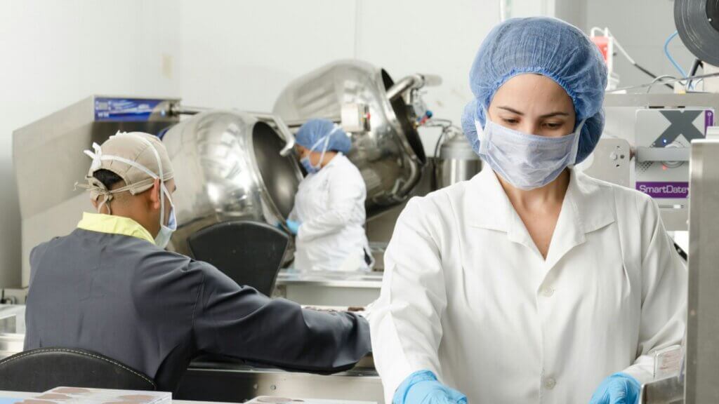 Three workers inside a factory wearing masks and protective coats