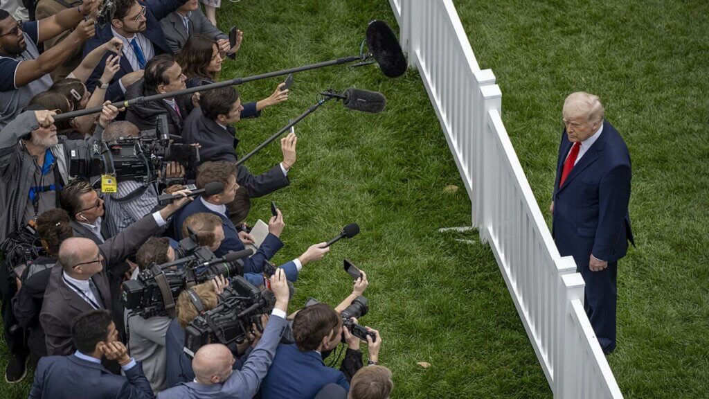 President Trump, in a suit, stands alone on the grass while a crowd of reporters and camera operators lean over a white fence with microphones and cameras pointed toward him.