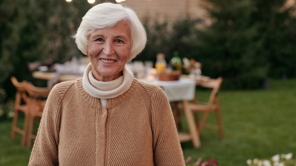 Senior woman with short hair smiling brightly while standing in a sunny backyard garden.