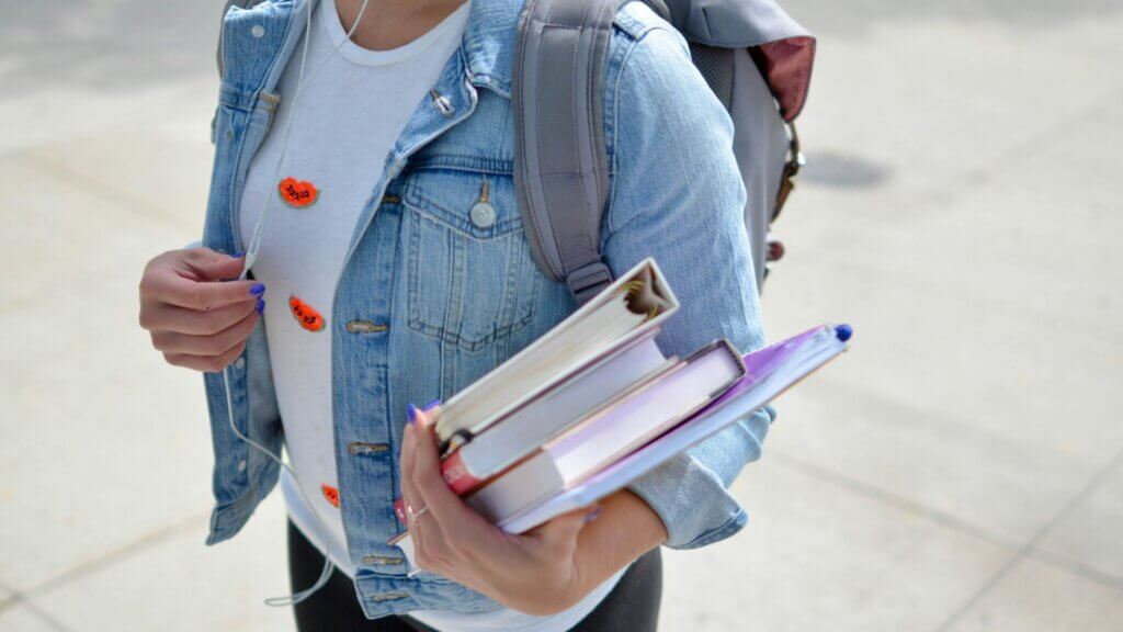College student carrying textbooks and wearing headphones on campus.