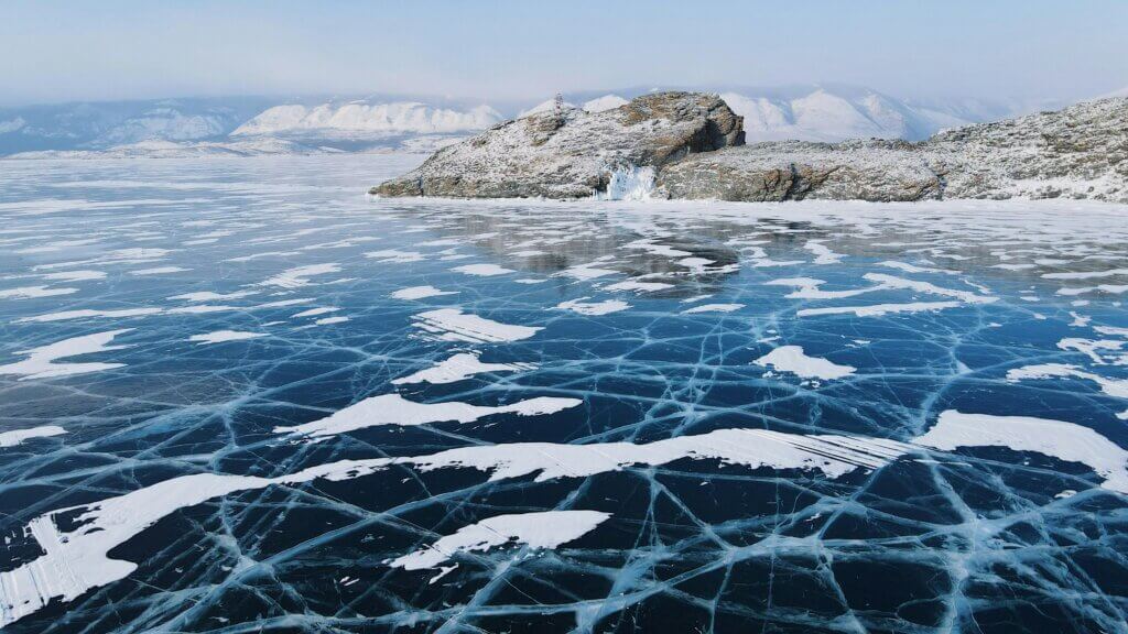 Frozen lake with large surface cracks near rocky mountains under a clear blue sky.