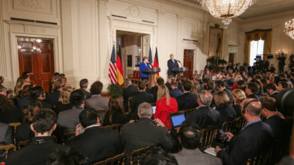 Press conference in a formal hall with U.S. and German flags behind two leaders addressing an audience.
